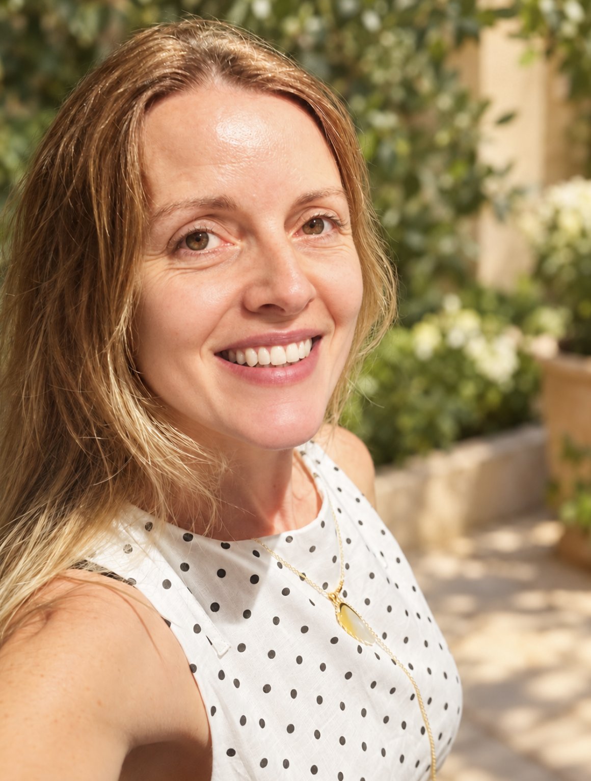 Núria smiling warmly in a sunlit garden, wearing a white polka-dot dress